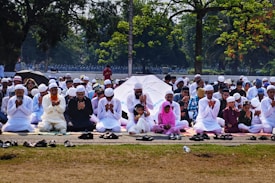 A large group of people are seated outdoors on mats, engaged in prayer. Most individuals are dressed in white clothing and wearing traditional caps. The setting is a park or open area surrounded by trees, with some people holding umbrellas for shade.