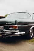 A vintage car with a shiny dark green exterior and chrome details is parked on a dirt path. The rear section of the vehicle is prominently visible, featuring a distinct emblem and dual exhaust pipes. The classic design suggests a focus on elegance and nostalgia.