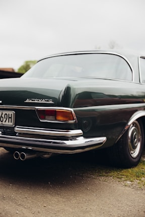 A vintage car with a shiny dark green exterior and chrome details is parked on a dirt path. The rear section of the vehicle is prominently visible, featuring a distinct emblem and dual exhaust pipes. The classic design suggests a focus on elegance and nostalgia.
