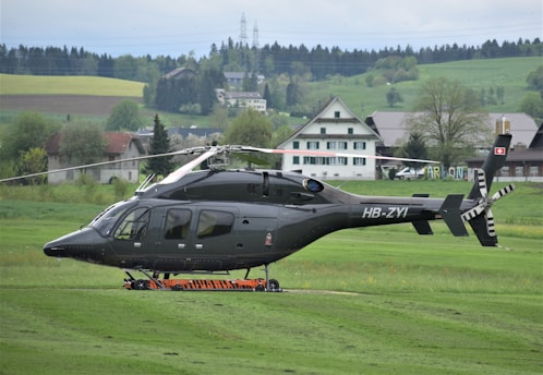 A sleek, dark-colored helicopter is parked on a grassy field, with a backdrop of rural houses and lush greenery. The atmosphere is calm and serene, showcasing a peaceful countryside setting.