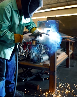A person wearing protective welding gear, including a helmet, gloves, and a green jacket, is engaged in a welding task. The welding process produces bright, blue-white sparks and a haze of smoke, illuminating the surrounding area. Various metal tools and equipment are visible in the slightly blurred background of the workshop.