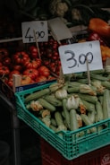 A market stall displaying a variety of fresh vegetables including a crate of zucchinis with blossoms and a pile of ripe red tomatoes. Two price tags are prominently displayed above the produce, indicating prices per kilogram. In the background, other vegetables like cauliflower and bell peppers are visible.