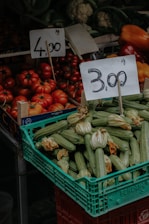 A market stall displaying a variety of fresh vegetables including a crate of zucchinis with blossoms and a pile of ripe red tomatoes. Two price tags are prominently displayed above the produce, indicating prices per kilogram. In the background, other vegetables like cauliflower and bell peppers are visible.