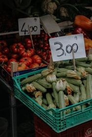A market stall displaying a variety of fresh vegetables including a crate of zucchinis with blossoms and a pile of ripe red tomatoes. Two price tags are prominently displayed above the produce, indicating prices per kilogram. In the background, other vegetables like cauliflower and bell peppers are visible.