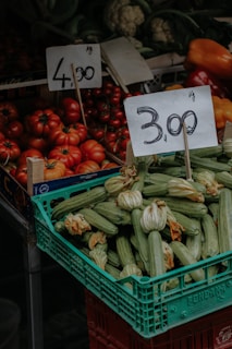 A market stall displaying a variety of fresh vegetables including a crate of zucchinis with blossoms and a pile of ripe red tomatoes. Two price tags are prominently displayed above the produce, indicating prices per kilogram. In the background, other vegetables like cauliflower and bell peppers are visible.
