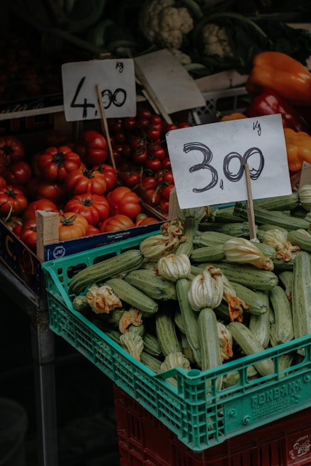 A market stall displaying a variety of fresh vegetables including a crate of zucchinis with blossoms and a pile of ripe red tomatoes. Two price tags are prominently displayed above the produce, indicating prices per kilogram. In the background, other vegetables like cauliflower and bell peppers are visible.