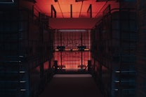 A dimly lit storage space features large, transparent plastic containers stacked on both sides, creating a narrow corridor. The room is illuminated by a warm red-orange light coming from above, with some graffiti visible on the containers' surfaces.