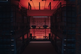 A dimly lit storage space features large, transparent plastic containers stacked on both sides, creating a narrow corridor. The room is illuminated by a warm red-orange light coming from above, with some graffiti visible on the containers' surfaces.