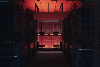 A dimly lit storage space features large, transparent plastic containers stacked on both sides, creating a narrow corridor. The room is illuminated by a warm red-orange light coming from above, with some graffiti visible on the containers' surfaces.
