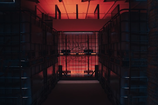 A dimly lit storage space features large, transparent plastic containers stacked on both sides, creating a narrow corridor. The room is illuminated by a warm red-orange light coming from above, with some graffiti visible on the containers' surfaces.