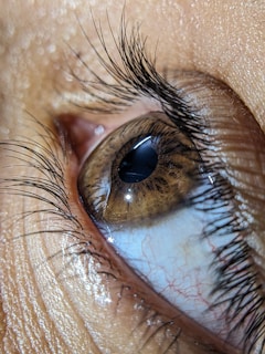 A detailed close-up of a human eye, showcasing the intricate patterns and details of the iris, the reflection on the cornea, and the surrounding eyelashes. The skin texture and tiny capillaries in the white of the eye are clearly visible.