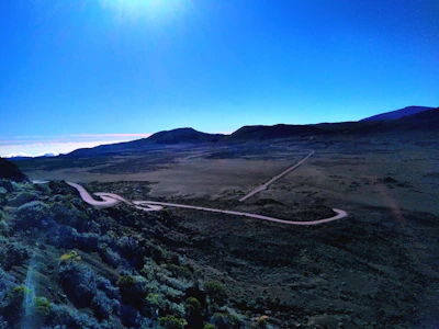 A winding dirt road cutting through vast, open outback plains under a bright blue sky.
