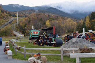A scenic landscape featuring vintage steam engines displayed in a park area, surrounded by autumn foliage. The scene is framed by a mountain range in the background partially covered by clouds. Two people are walking on a pathway bordered by flowers in pots and pumpkins. An old railway track ascends the hillside.
