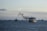 A large yellow ship emits black smoke as it sails on the sea. Several other ships are visible in the background, with a vast expanse of water surrounding them. The sky is overcast with a few clouds, giving the scene a somewhat somber atmosphere.
