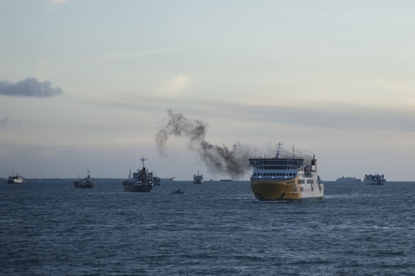 A large yellow ship emits black smoke as it sails on the sea. Several other ships are visible in the background, with a vast expanse of water surrounding them. The sky is overcast with a few clouds, giving the scene a somewhat somber atmosphere.