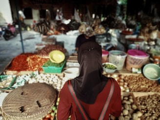 A person wearing a dark headscarf stands in front of a market stall filled with an array of fresh produce. The stall has baskets of potatoes, chili peppers, garlic, and various other vegetables, with plastic containers and bowls placed among the goods. The background shows a blurred view of more market items and another person attending to the stall.