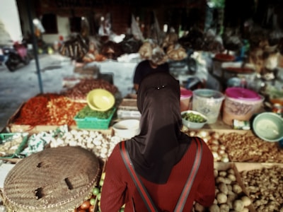 A person wearing a dark headscarf stands in front of a market stall filled with an array of fresh produce. The stall has baskets of potatoes, chili peppers, garlic, and various other vegetables, with plastic containers and bowls placed among the goods. The background shows a blurred view of more market items and another person attending to the stall.