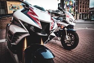 Two sport motorcycles parked on a brick-paved street. The foreground motorcycle features white, red, and dark graphics with detailed front headlights. In the background, another bike is visible with similar sporty decals. The setting includes an urban backdrop with a colorful mural on a building.