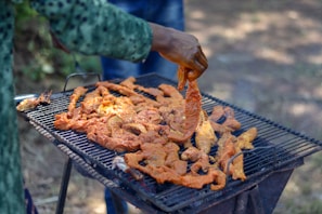 A person is grilling marinated meat on a barbecue grill, using their hand to turn or arrange the pieces. The grill is positioned outdoors and is filled with various cuts of meat that appear to be seasoned.