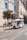 A small, white motorized tuk-tuk with several passengers is driving along a cobblestone street. The street is lined with old buildings, featuring colorful blue window shutters. A tree with foliage stands next to a green bench, and a pedestrian is walking nearby.