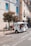 A small, white motorized tuk-tuk with several passengers is driving along a cobblestone street. The street is lined with old buildings, featuring colorful blue window shutters. A tree with foliage stands next to a green bench, and a pedestrian is walking nearby.