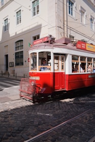 A vintage red tram is moving along cobblestone tracks in a city setting. The tram is adorned with signage related to tours and tickets. Sunlight casts shadows on the building facades and the street. Pedestrians are visible inside the tram, enjoying the ride through the city.