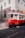 A vintage red tram is moving along cobblestone tracks in a city setting. The tram is adorned with signage related to tours and tickets. Sunlight casts shadows on the building facades and the street. Pedestrians are visible inside the tram, enjoying the ride through the city.
