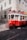 A vintage red tram is moving along cobblestone tracks in a city setting. The tram is adorned with signage related to tours and tickets. Sunlight casts shadows on the building facades and the street. Pedestrians are visible inside the tram, enjoying the ride through the city.