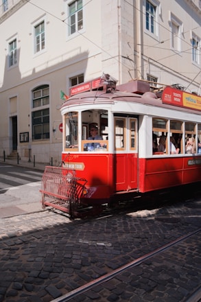 A vintage red tram is moving along cobblestone tracks in a city setting. The tram is adorned with signage related to tours and tickets. Sunlight casts shadows on the building facades and the street. Pedestrians are visible inside the tram, enjoying the ride through the city.