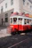 A vintage red tram is moving along cobblestone tracks in a city setting. The tram is adorned with signage related to tours and tickets. Sunlight casts shadows on the building facades and the street. Pedestrians are visible inside the tram, enjoying the ride through the city.