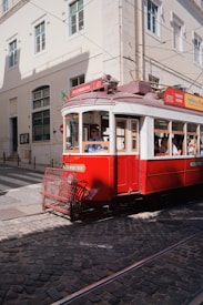 A vintage red tram is moving along cobblestone tracks in a city setting. The tram is adorned with signage related to tours and tickets. Sunlight casts shadows on the building facades and the street. Pedestrians are visible inside the tram, enjoying the ride through the city.