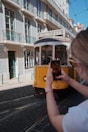 A historic yellow tram travels down a cobblestone street lined with traditional tiled buildings. A person in the foreground captures the scene on a smartphone, highlighting the quaint architecture and vibrant street life.