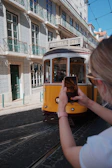 A historic yellow tram travels down a cobblestone street lined with traditional tiled buildings. A person in the foreground captures the scene on a smartphone, highlighting the quaint architecture and vibrant street life.