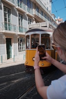 A historic yellow tram travels down a cobblestone street lined with traditional tiled buildings. A person in the foreground captures the scene on a smartphone, highlighting the quaint architecture and vibrant street life.