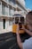 A historic yellow tram travels down a cobblestone street lined with traditional tiled buildings. A person in the foreground captures the scene on a smartphone, highlighting the quaint architecture and vibrant street life.