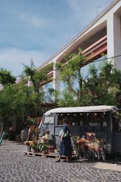 A woman stands in front of a vintage van that has been converted into a flower shop. Various potted plants and bouquets are displayed on stands and wooden pallets in front of the van. The setting is outdoors on a cobblestone surface with a large overpass above and trees surrounding the area. The sky is clear and sunny, adding a vibrant feel to the scene.