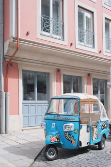 A vibrantly painted blue tuk-tuk with floral and heart motifs is parked on a cobblestone street next to a pastel pink building with large windows and decorative railings.