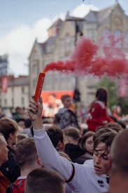 A crowd of people gathers in a lively outdoor setting, with many wearing sports jerseys. A young person in the foreground holds up a red smoke flare, which releases a cloud of red smoke. Background buildings include classical architecture.