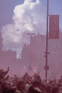 A large crowd of people gathers on a city street, with many holding up their phones. Smoke fills the air, colored pinkish-red, suggesting a festive or celebratory atmosphere. Buildings with classic architecture and a Premier League banner are visible in the background under a partly cloudy sky.
