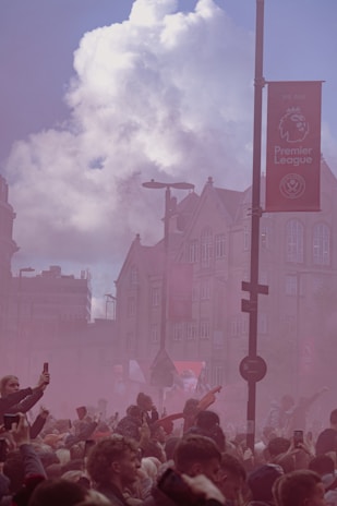A large crowd of people gathers on a city street, with many holding up their phones. Smoke fills the air, colored pinkish-red, suggesting a festive or celebratory atmosphere. Buildings with classic architecture and a Premier League banner are visible in the background under a partly cloudy sky.