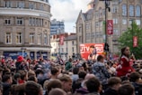 A large crowd of people, many wearing red and white, gather in a city square. They are watching an event broadcast on a large screen, which displays a man speaking into a microphone with logos suggesting a sporting event. Surrounding buildings are historical and commercial, with banners indicating a connection to the Premier League.