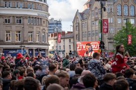 A large crowd of people, many wearing red and white, gather in a city square. They are watching an event broadcast on a large screen, which displays a man speaking into a microphone with logos suggesting a sporting event. Surrounding buildings are historical and commercial, with banners indicating a connection to the Premier League.