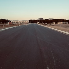 A newly paved road extends into the distance flanked by areas of dirt and sparse vegetation. At the far end, there is a row of buildings with a mix of trees scattered around. The sky is clear, giving a hint of evening light.