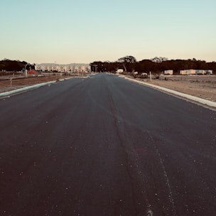 A newly paved road extends into the distance flanked by areas of dirt and sparse vegetation. At the far end, there is a row of buildings with a mix of trees scattered around. The sky is clear, giving a hint of evening light.