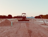 A bulldozer faces forward on a partially developed construction site with mounds of dirt on both sides. The site is relatively barren, with sparse vegetation and trees visible in the background. There is a blue building in the midground, and the sky appears clear with a soft gradient from light blue to a pale hue.