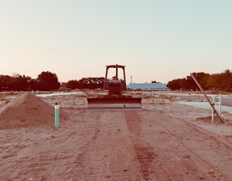 A bulldozer faces forward on a partially developed construction site with mounds of dirt on both sides. The site is relatively barren, with sparse vegetation and trees visible in the background. There is a blue building in the midground, and the sky appears clear with a soft gradient from light blue to a pale hue.