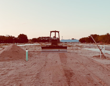 A bulldozer faces forward on a partially developed construction site with mounds of dirt on both sides. The site is relatively barren, with sparse vegetation and trees visible in the background. There is a blue building in the midground, and the sky appears clear with a soft gradient from light blue to a pale hue.
