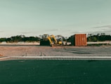A construction site with a yellow excavator and a container on a dirt field, surrounded by trees in the distance under a clear sky.
