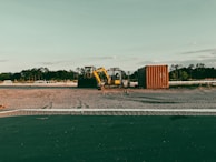 Wide shot of an excavator clearing land surrounded by trees and blue sky.