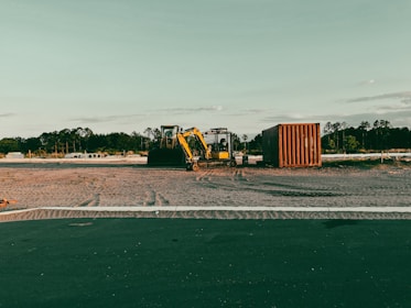 A construction site with a yellow excavator and a container on a dirt field, surrounded by trees in the distance under a clear sky.